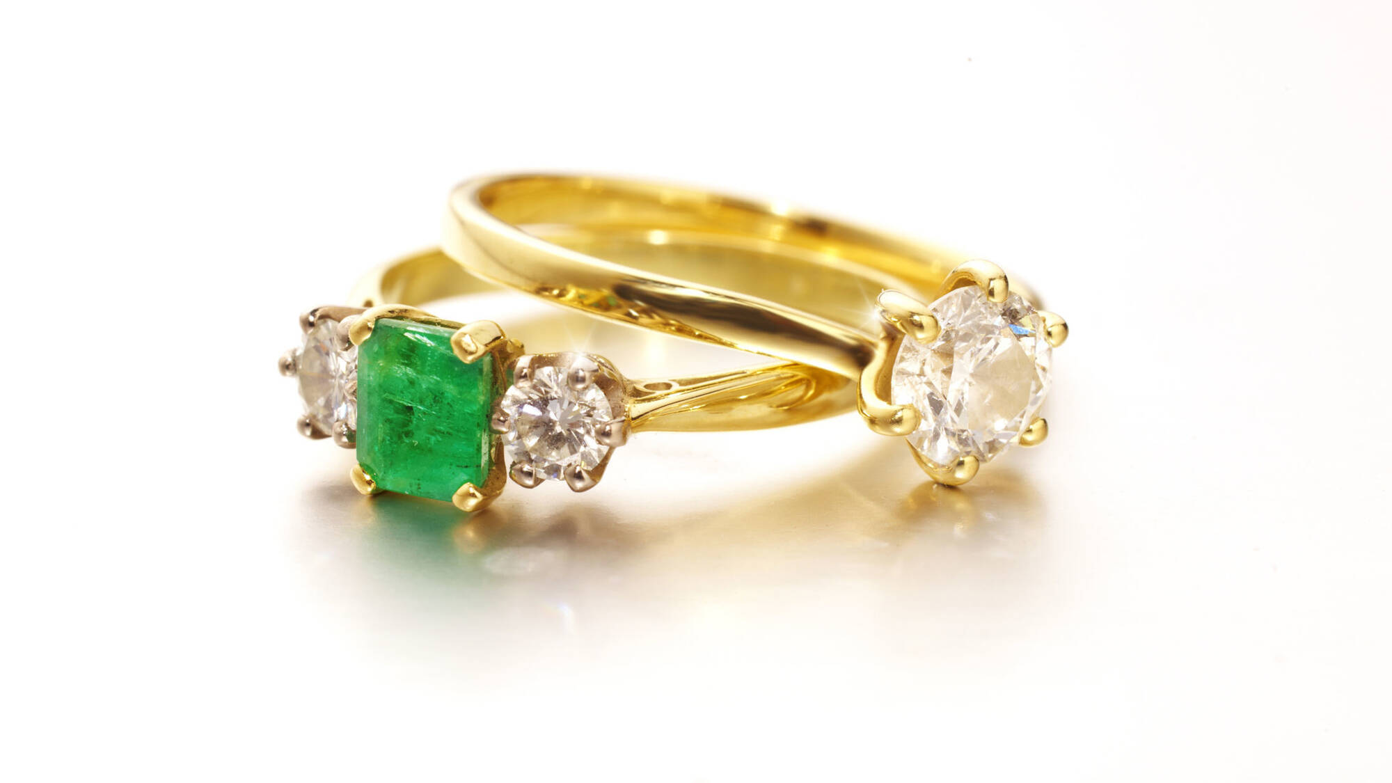 A matching pair of handcrafted gold rings featuring a vibrant emerald cut stone, diamond accents, and a classic solitaire engagement ring.