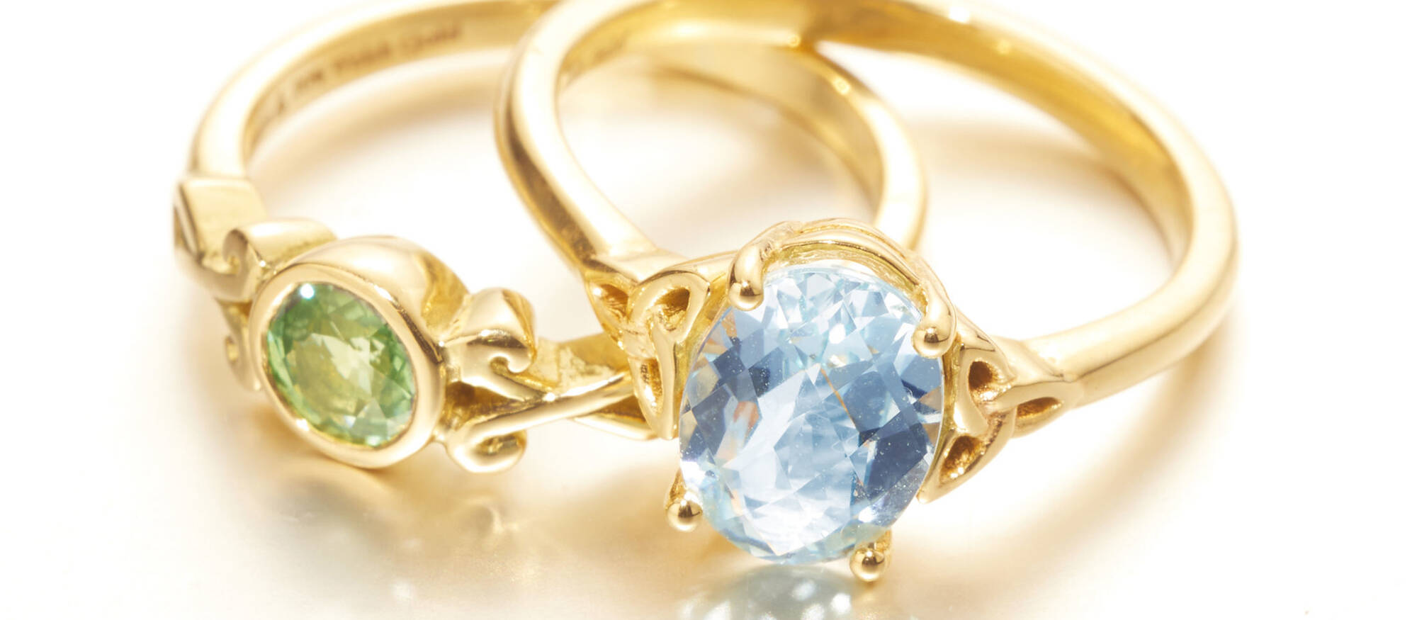 Two elegant gold rings, one with a prong-set oval blue aquamarine and one with a round green gemstone, illustrating the range of bespoke Welsh gold jewellery.