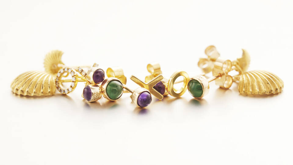 Close-up of handcrafted gold jewelry including diamond-set circular studs, vibrant gemstone cabochons, and intricately detailed shell-shaped earrings.
