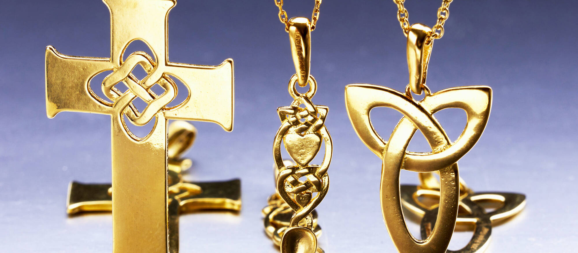A collection of three solid Welsh gold necklaces featuring a large Celtic cross with knotwork, a traditional Welsh lovespoon pendant with heart details, and a modern interlocking triquetra symbol.