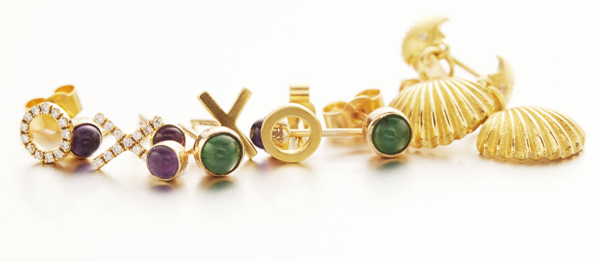 A close-up of several small gold earrings arranged in a row, showcasing a mix of purple and green gemstones alongside symbolic shapes and textured sea shell designs in high-polish gold.