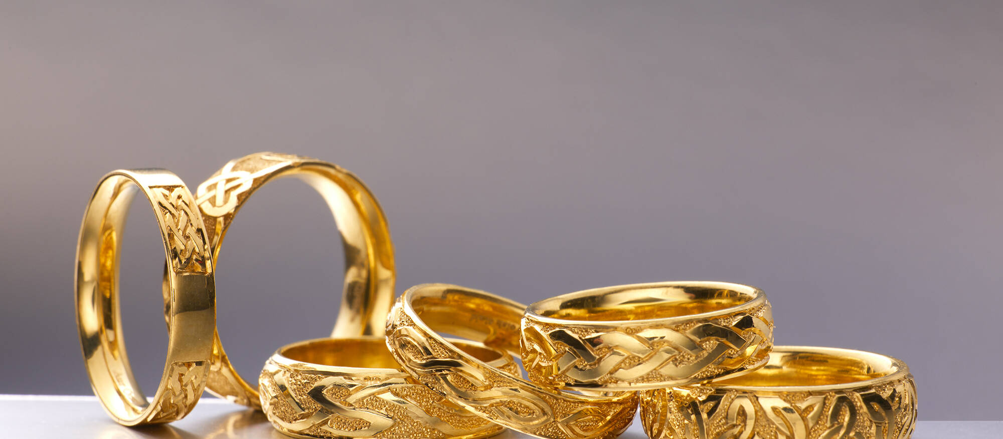 An editorial display of several Welsh gold bands with various symbolic Celtic engravings and interlocking patterns, arranged on a reflective surface.