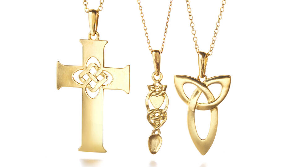 Close-up of handcrafted gold necklaces featuring a polished cross, an intricate lovespoon with heart detail, and a smooth-edged Celtic knot pendant.