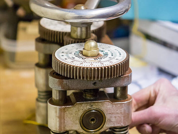 An action shot where a jeweller's hand is visible operating a mechanical rolling mill to achieve the precise thickness required for a handcrafted gold ring.