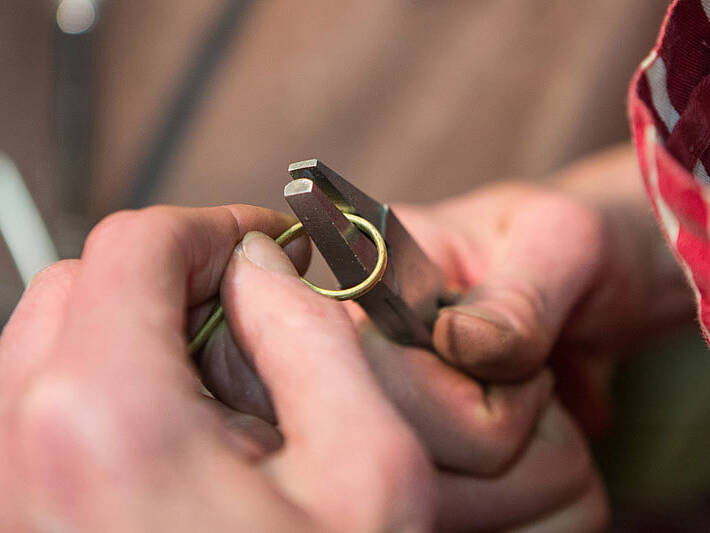 A close-up action shot showing a jeweller's hands using heavy-duty parallel pliers to carefully bend a length of Welsh gold into a circular ring shape.