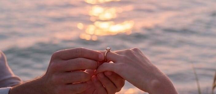 A close-up, lifestyle shot of a marriage proposal at sunset on a sandy beach, showing a person's hands placing a gold diamond engagement ring onto their partner's finger with the ocean waves in the background.