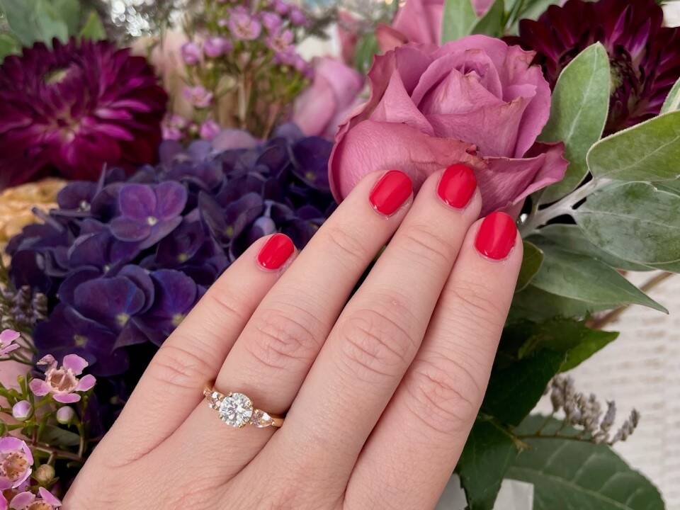 Close-up of an image featuring a round brilliant-cut centre diamond flanked by two smaller diamonds on a polished Welsh gold band, worn by Charlotte with a vibrant red manicure.