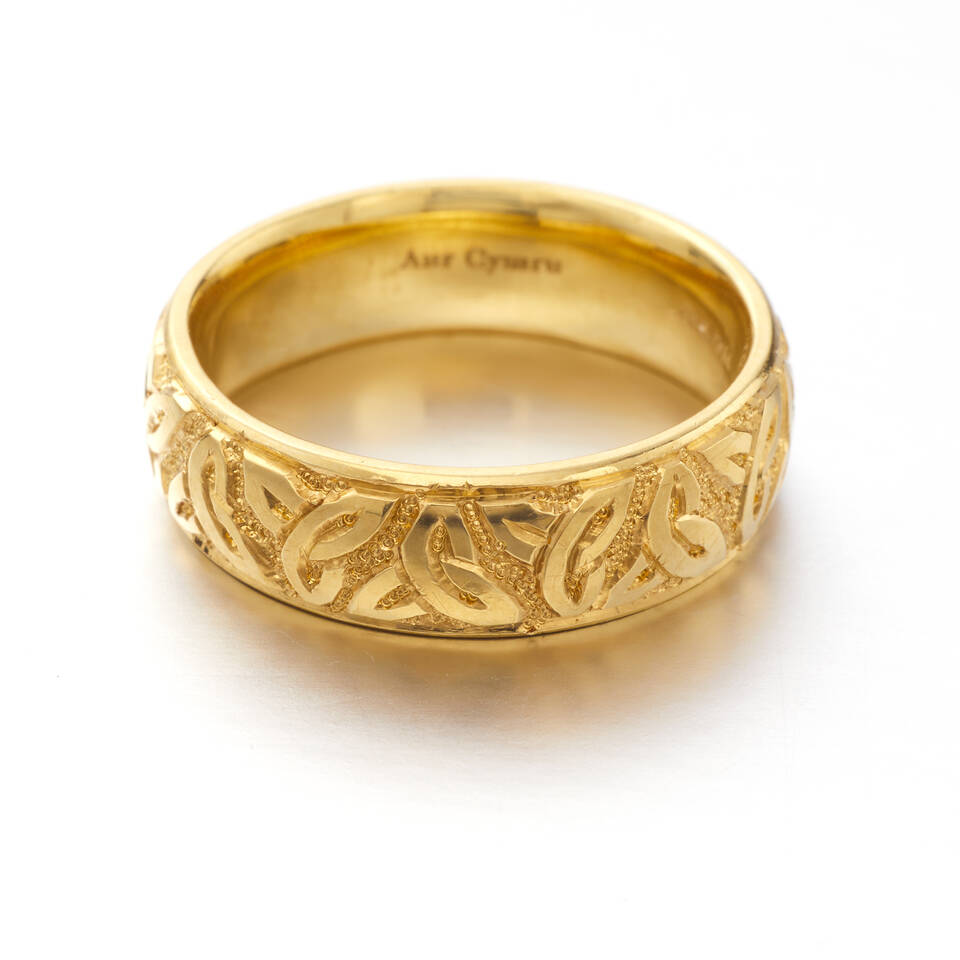 A detailed studio photograph of a wide gold wedding ring featuring a raised Celtic knot pattern and a textured background, highlighting traditional Welsh artistry.