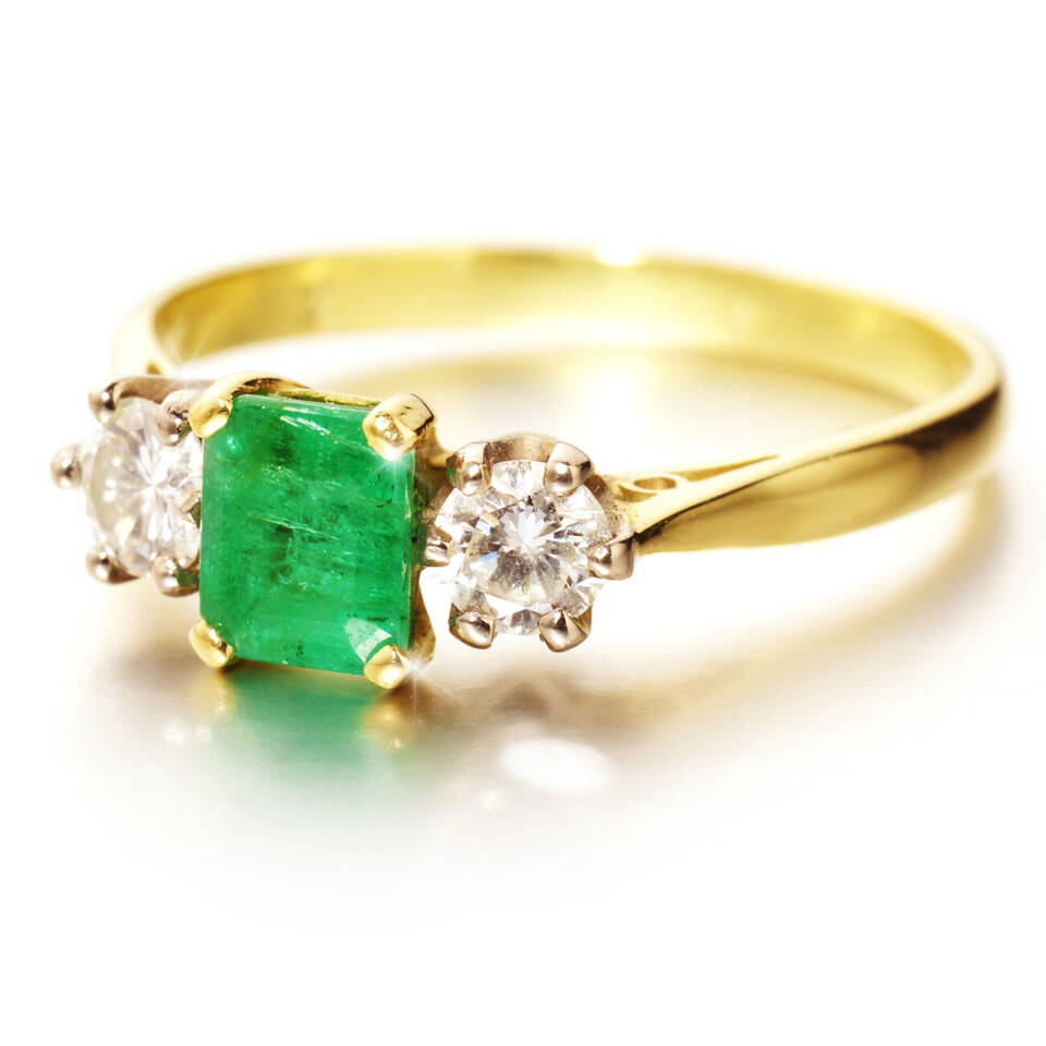 Close-up of a handcrafted gold trilogy ring showcasing a vibrant green emerald and sparkling diamond accents on a polished gold band.
